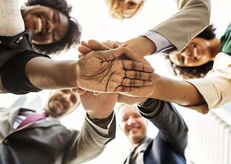 Zu sehen ist eine im Kreis stehende Gruppe Menschen unterschiedlichen Alters und Geschlechts sowie verschiedener Hautfarben. Sie sind aus der Froschperspektive aufgenommen. Ihre Hände liegen übereinander in der Bildmitte und die Menschen lachen in die Kamera.
