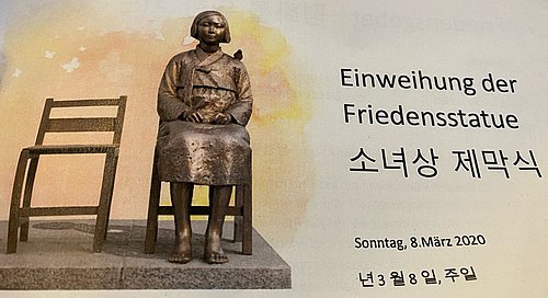 Auf dem Bild ist ein leerer Bronzestuhl und daneben die auf einem Stuhl sitzende Statue einer jungen Frau