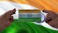 Das Bild zeigt zwei Hände, die eine OP-Maske in den indischen Nationalfarben halten. Im Hintergrund weht die indische Flagge.