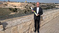 Ein Mann im Anzug steht auf einem gepflasterten Weg neben einer Steinmauer, hinter der sich das Panorama Jerusalems erstreckt. Links im Hintergrund sind markante Gebäude wie der Felsendom mit goldener Kuppel und die al-Aqsa-Moschee zu erkennen. Die Kerze zeigt Motive in Blau, Gelb und Weiß.